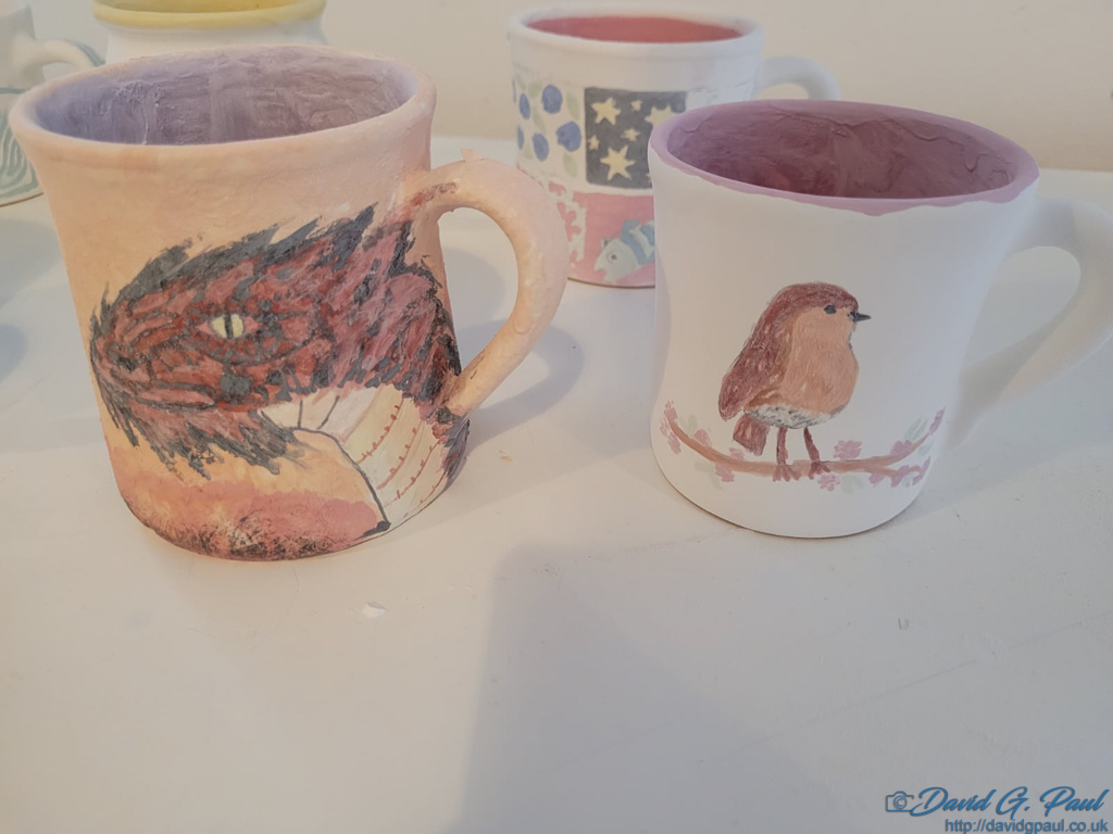 An orange mug with a dragon painted on it, besides a white mug with a robin painted on