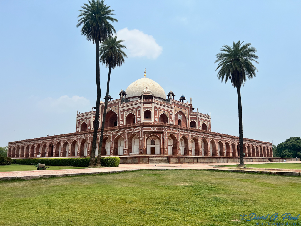 A red sandstone and white marble building on two tiers, with a white dome. There are palm trees in front of this with cultivated green grass.