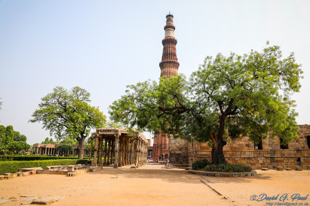A very tall minaret towers over ruined buildings and trees