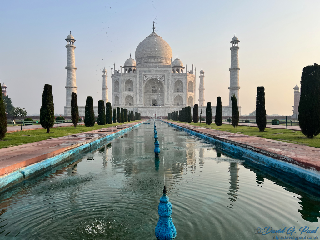 The Taj Mahal at dawn with a pool of water and blue water fountains in front. There are conical trees either side of the water.