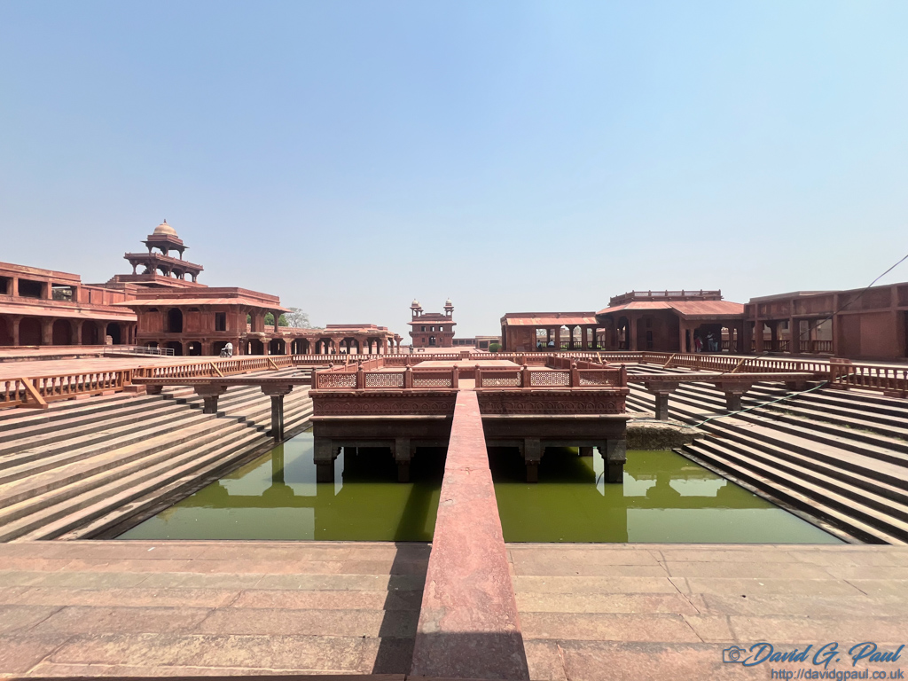 A path leading to a centre platform surrounded by water. Numerous red sandstone buildings surround the steps down to the water.