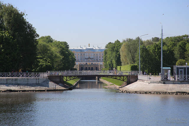 A canal with a bridge across it, which leads to a large house. Trees line the canal