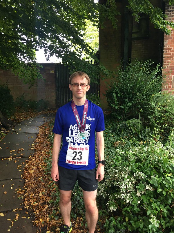 Caucasian male standing in front of a bush wearing black shorts and a blue t-shirt with a race number attached, and a medal hanging from his neck