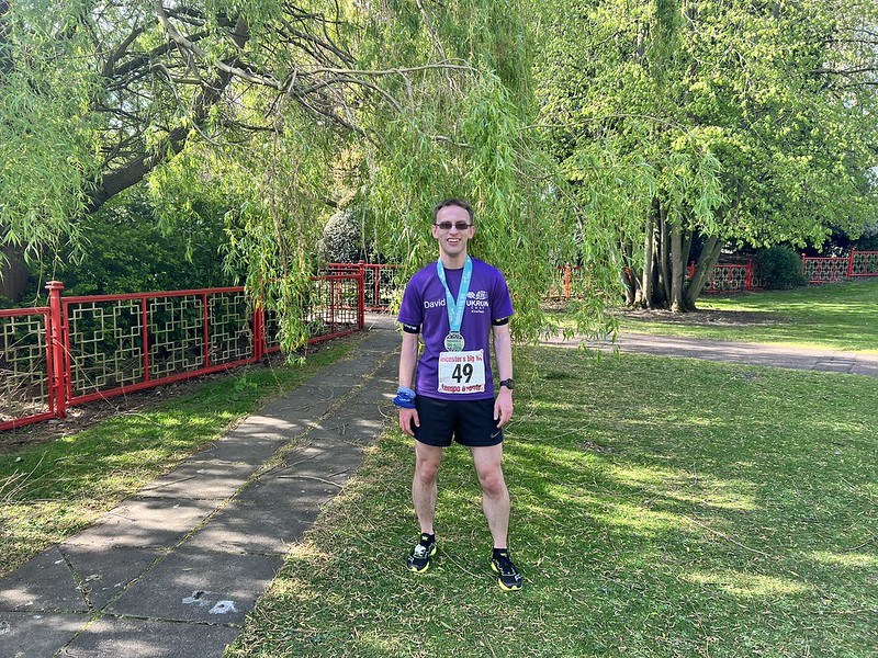 A male runner wearing a purple t-shirt and black shorts and sunglasses. He has a medal around his neck also, and is standing on grass with trees and a red fence behind