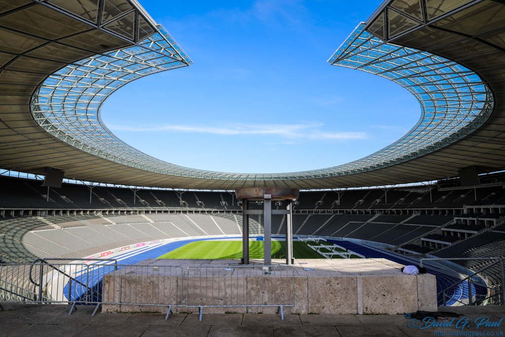 Olympic Flame Bowl, and beyond you can see seating, a blue running track around  afield, and above is a roof with a cutout above the bowl