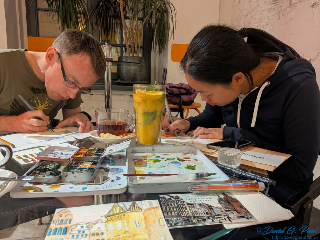 Two people busy painting with watercolours on a table full of paper, and paints